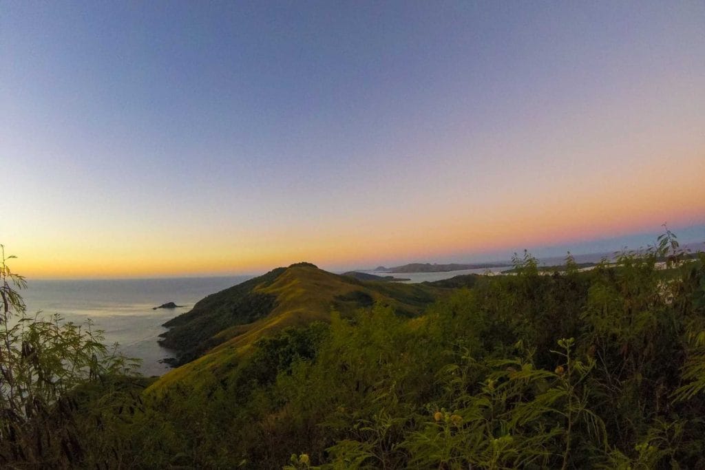 Sunrise over rolling green hills by the ocean, viewed from a high vantage point surrounded by trees.