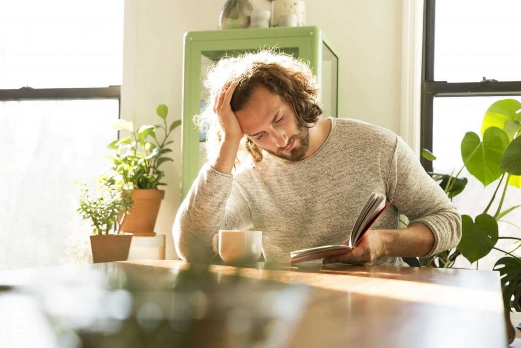 Man reading a book in a bright room with natural light and houseplants, creating a calm and mindful atmosphere.