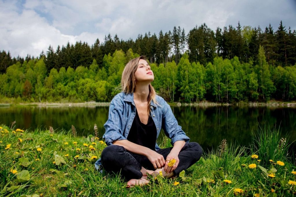 Woman sitting cross-legged in a meadow beside a lake, eyes closed and face tilted toward the sky, symbolising body awareness and connection with nature.