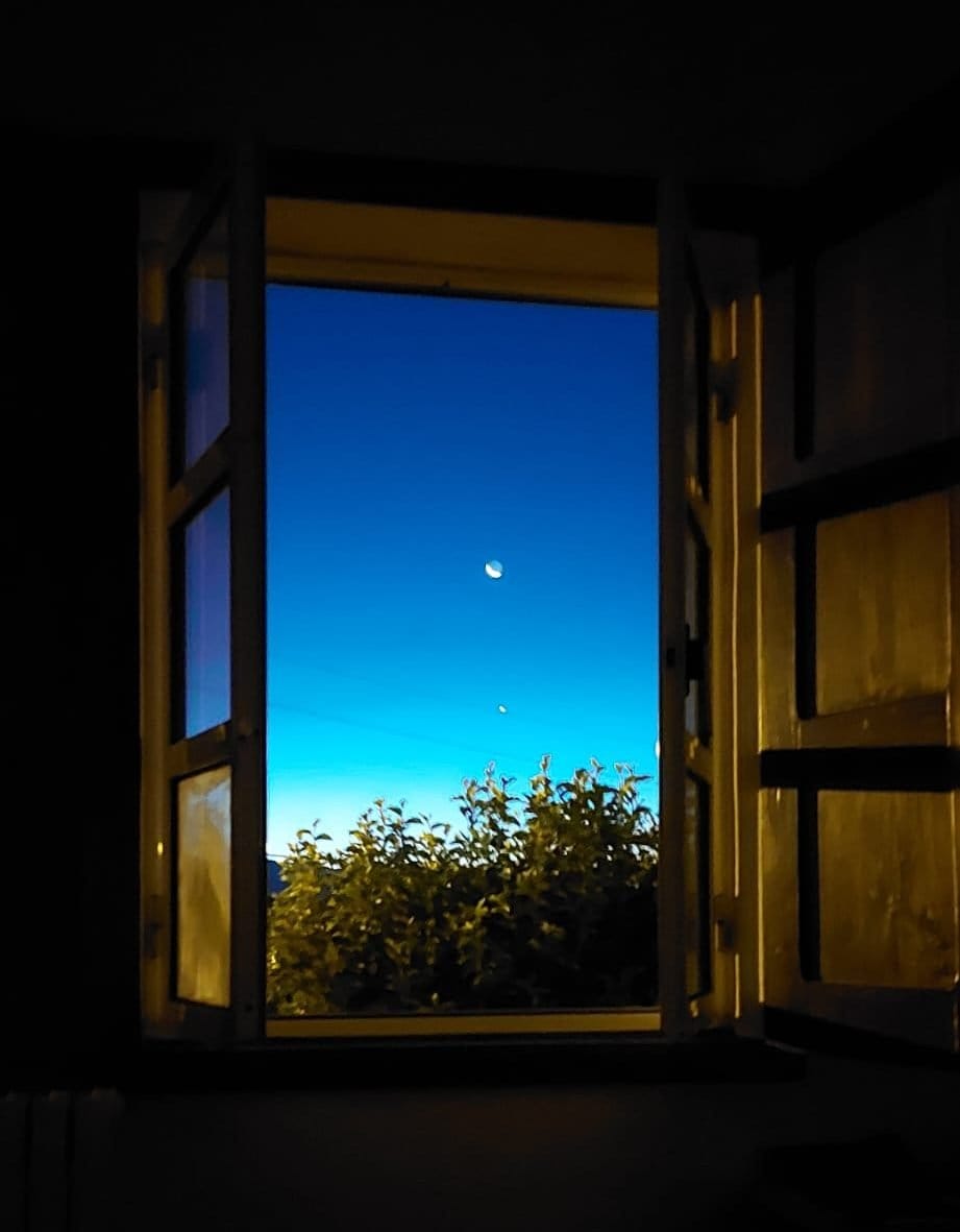 Open window at night showing a crescent moon over trees, symbolising reflection and calm.